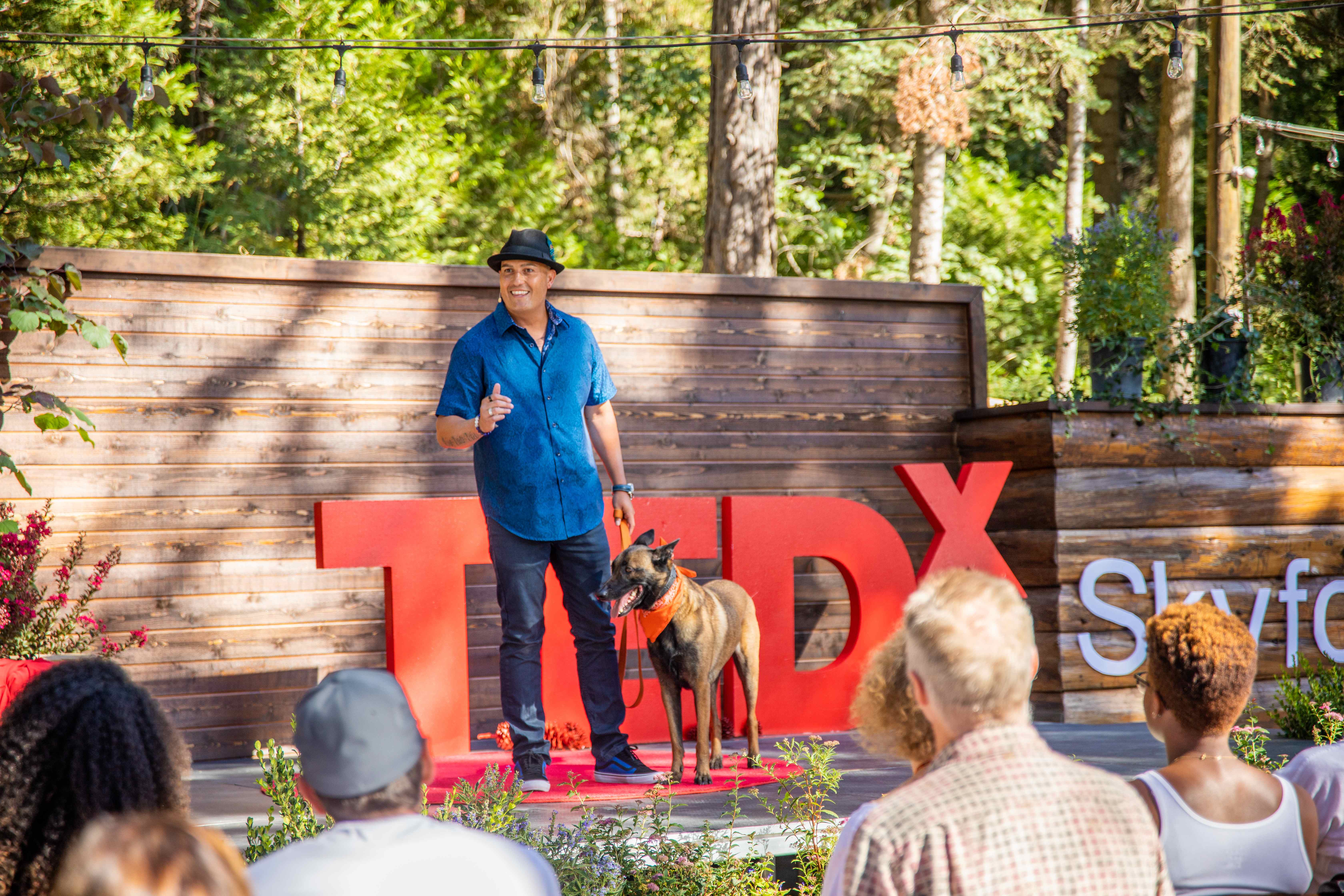 Ryan Matthews speaking on stage at TEDx Skyforest with his Belgian Malinois beside him